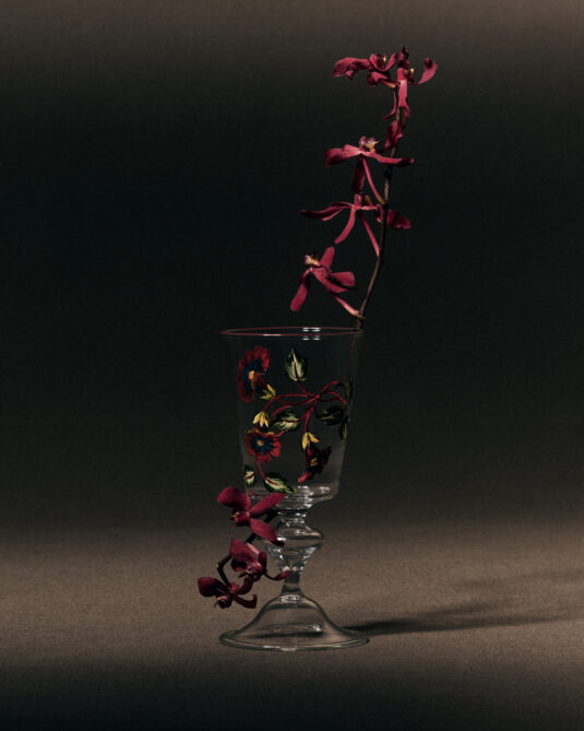 Un verre à eau transparent de la collection Ranelagh de Dior Maison, peint à la main avec un délicat motif floral rouge et jaune, une fleur séchée pourpre est drapée autour du verre