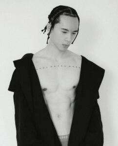 Portrait de Ritchie Tran Ve, torse nu, portant un manteau noir ouvert. Il pose face à l’objectif avec douceur, tresses plaquées, éclairage doux minimaliste. Photographie d’Elys Berroterán pour la cover story SPACE SONG de Salut les Garçons Magazine. Thom Browne & Skims.