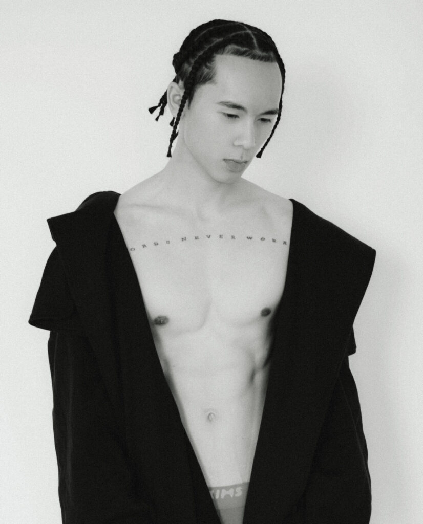 Portrait de Ritchie Tran Ve, torse nu, portant un manteau noir ouvert. Il pose face à l’objectif avec douceur, tresses plaquées, éclairage doux minimaliste. Photographie d’Elys Berroterán pour la cover story SPACE SONG de Salut les Garçons Magazine. Thom Browne & Skims.