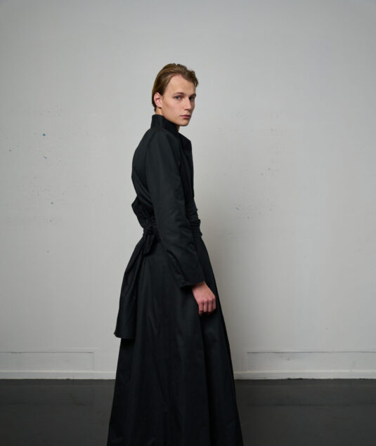 Silhouette KML FW26/27 dans l’espace bétonné de l’Institut du Monde Arabe : mannequin immobile puis en mouvement, vêtement long et fluide dans des tons sable et noir, éclairé par les reflets d’eau projetés sur les murs