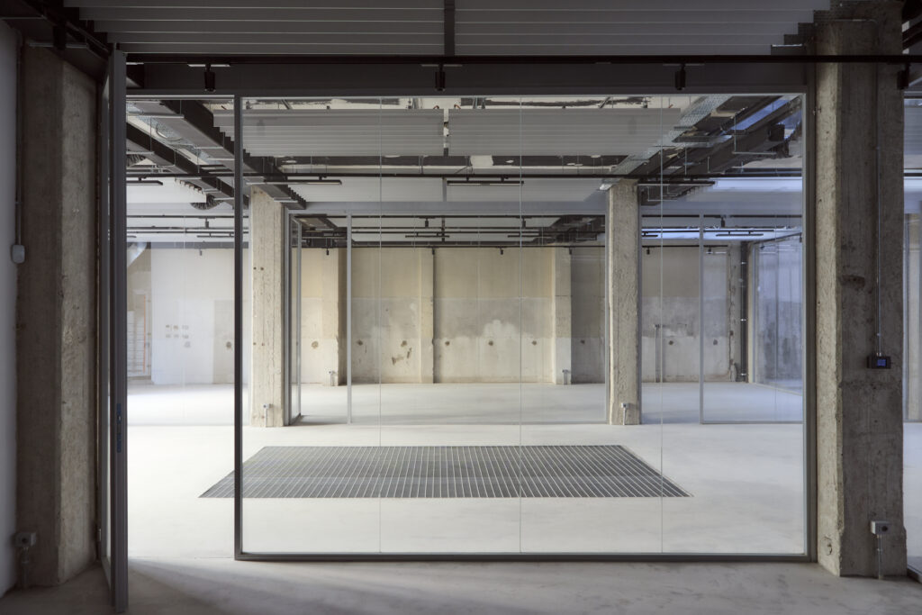 Interior of an empty industrial space with glass partition walls, concrete columns, and exposed ceiling plumbing/pipes on a bright, open floor.