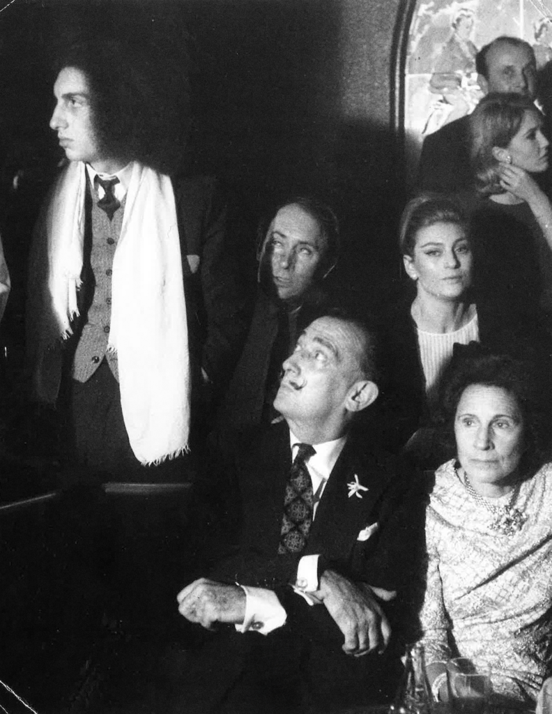 Black-and-white photo of a formal party: a tall man with a white scarf stands left while a seated man with a mustache and a woman sit nearby, looking upward or aside.