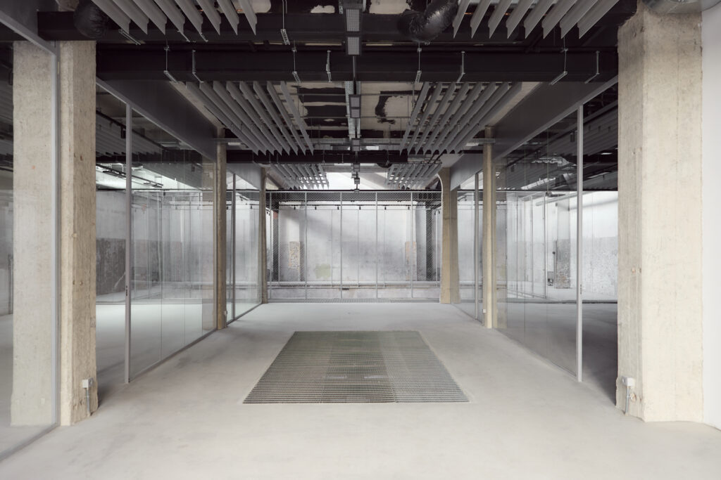 Industrial-style office corridor with glass walls and exposed ceiling, concrete columns, and a central metal floor grate under construction