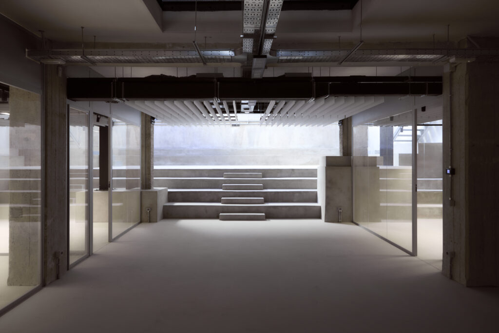Industrial interior corridor with glass walls on both sides and concrete steps leading up outside at the far end.