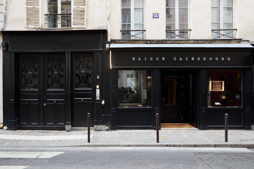 Paris storefront with black facade reading 'MAISON GAINSBOURG', ornate iron doors, and tall windows above.