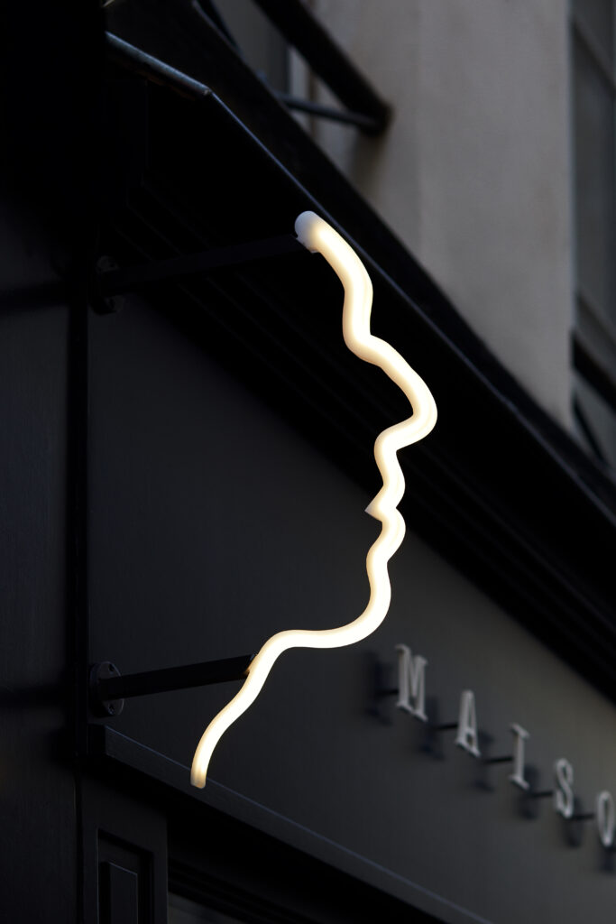 Glowing zigzag neon tube mounted on a dark exterior wall, with blurred letters in the background.