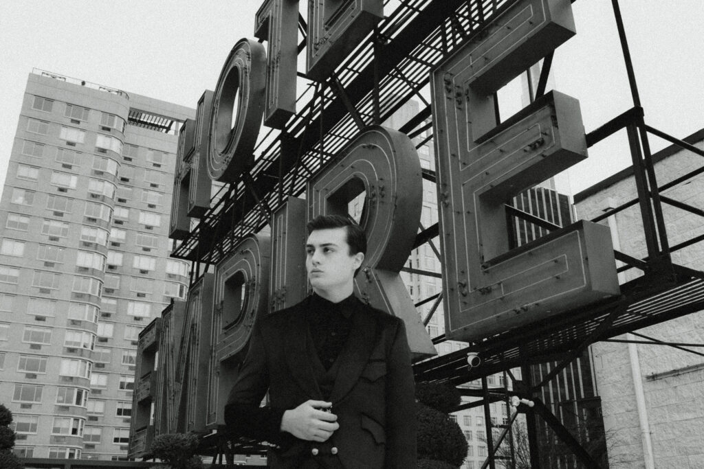Young man in a dark suit standing in front of a large metal sign structure with block letters and city buildings in the background.