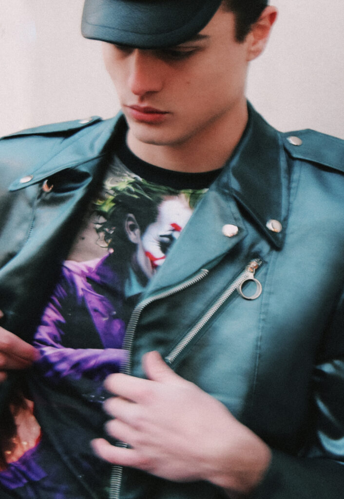 Young man in a teal leather jacket and cap, looking down while adjusting a colorful printed T-shirt underneath.