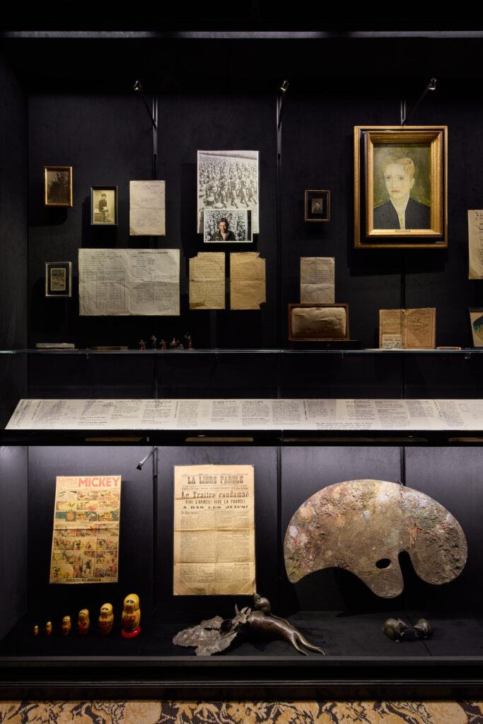Museum display wall with framed portraits, old documents, and a large gold-framed painting of a woman on a dark backdrop.