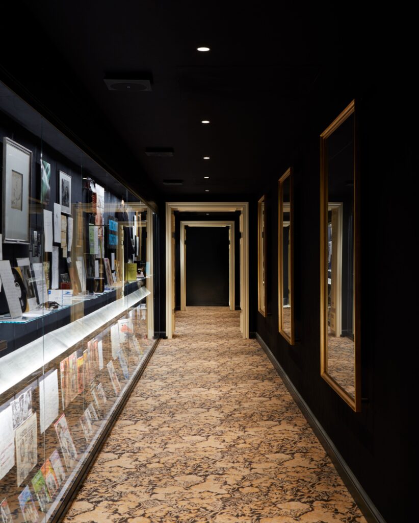 Dim museum hallway with glass display cases along the left and gold-framed wall panels on the right, leading to a dark doorway at the far end.