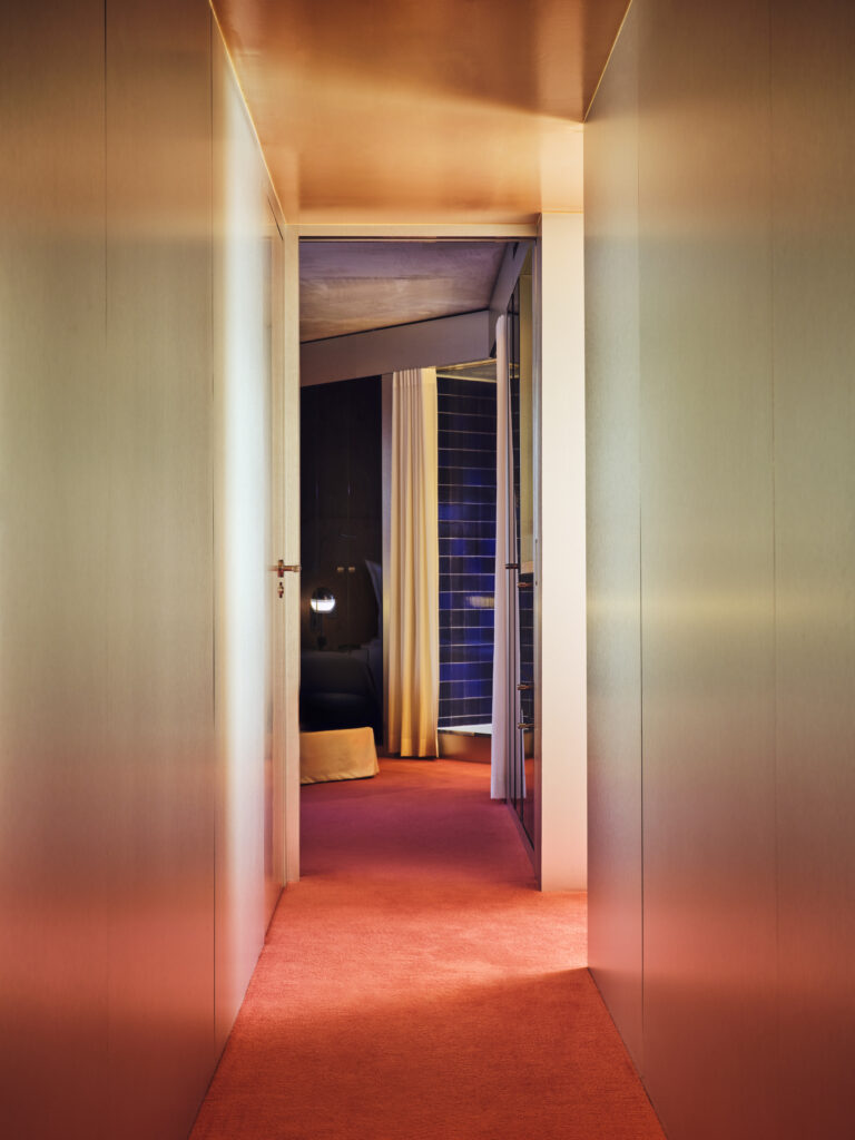Hotel hallway with orange carpet opens into a bedroom with a blue-tiled wall and beige curtains.