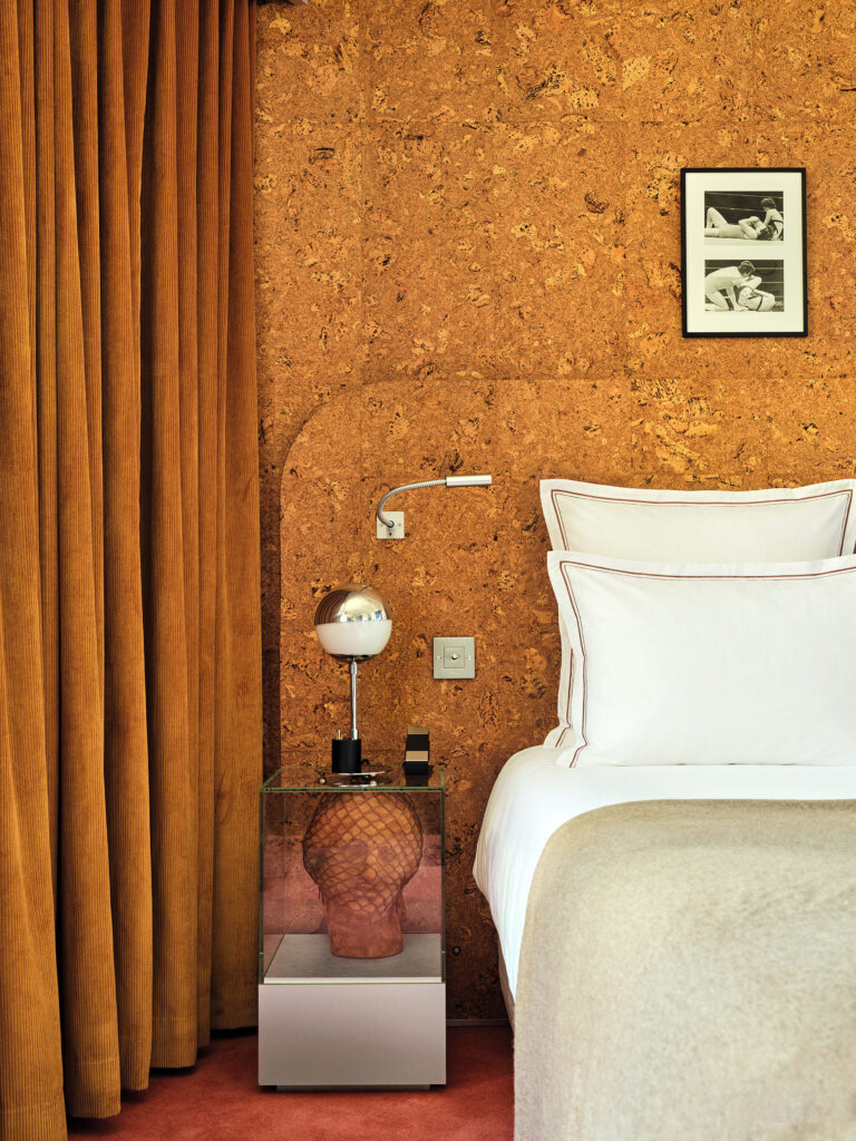 Bedroom corner with cork wall, brown curtains, and a neatly made bed with white pillows and a beige throw. A glass-topped nightstand holds a modern spherical lamp and a small black device; a ceramic bust sits inside the glass display beneath. Framed black-and-white photos hang on the wall above the bed.