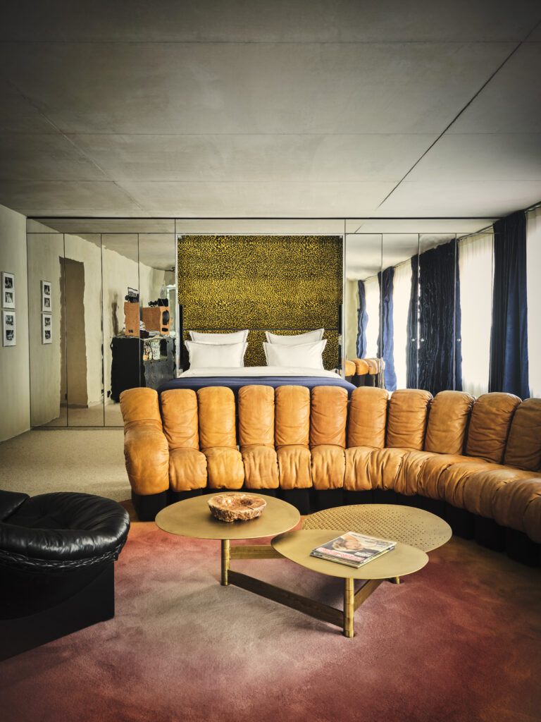 Living room with an orange tufted sectional, round gold coffee tables, and mirrored walls; leopard-print headboard behind a bed in the background.”] ,