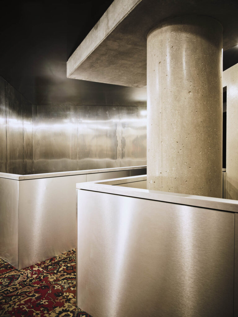 Modern stainless steel interior with a large concrete column and metal panels, on a patterned carpeted floor, suggesting an industrial lobby area.