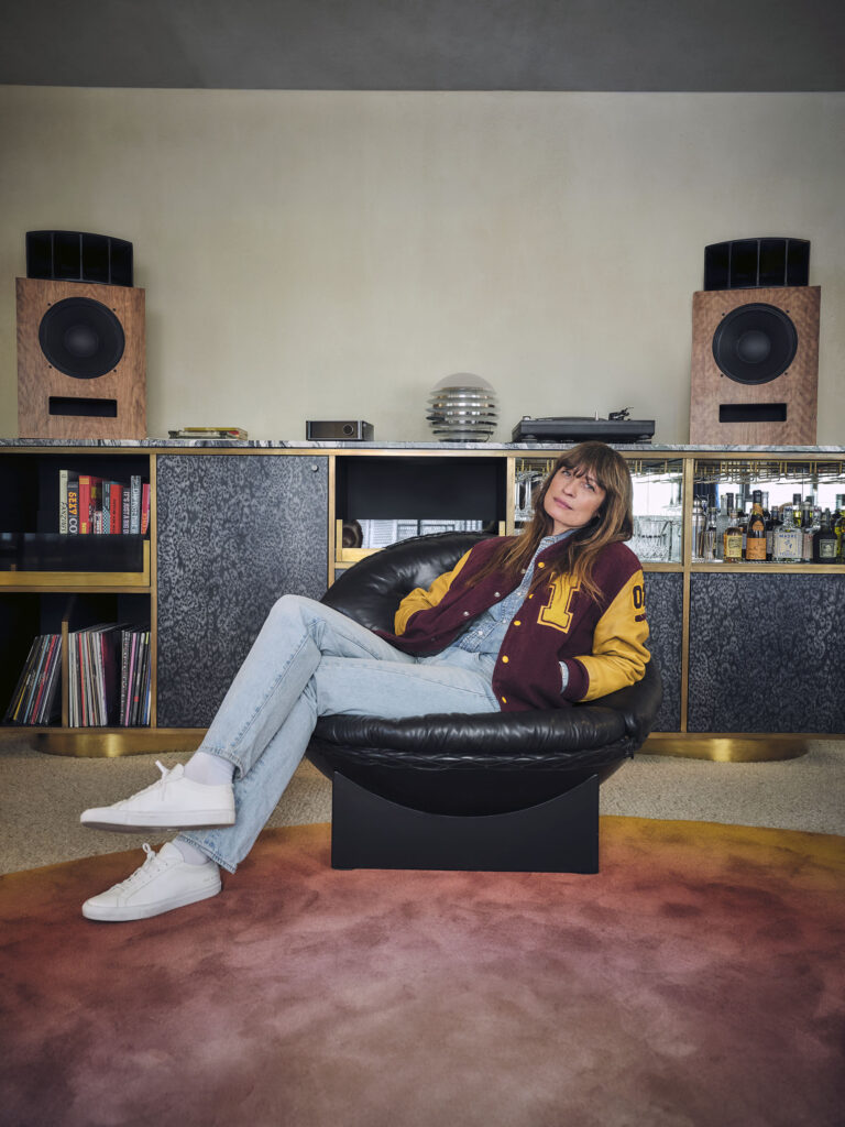 Woman relaxing in a retro-styled living room, sitting in a black round chair with a maroon-and-gold varsity jacket and light jeans. On shelves behind her are large wooden speakers and a bar area.