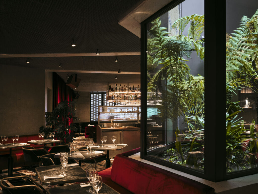 Restaurant dining area with tables set for guests and a glass-walled indoor garden visible to the right.