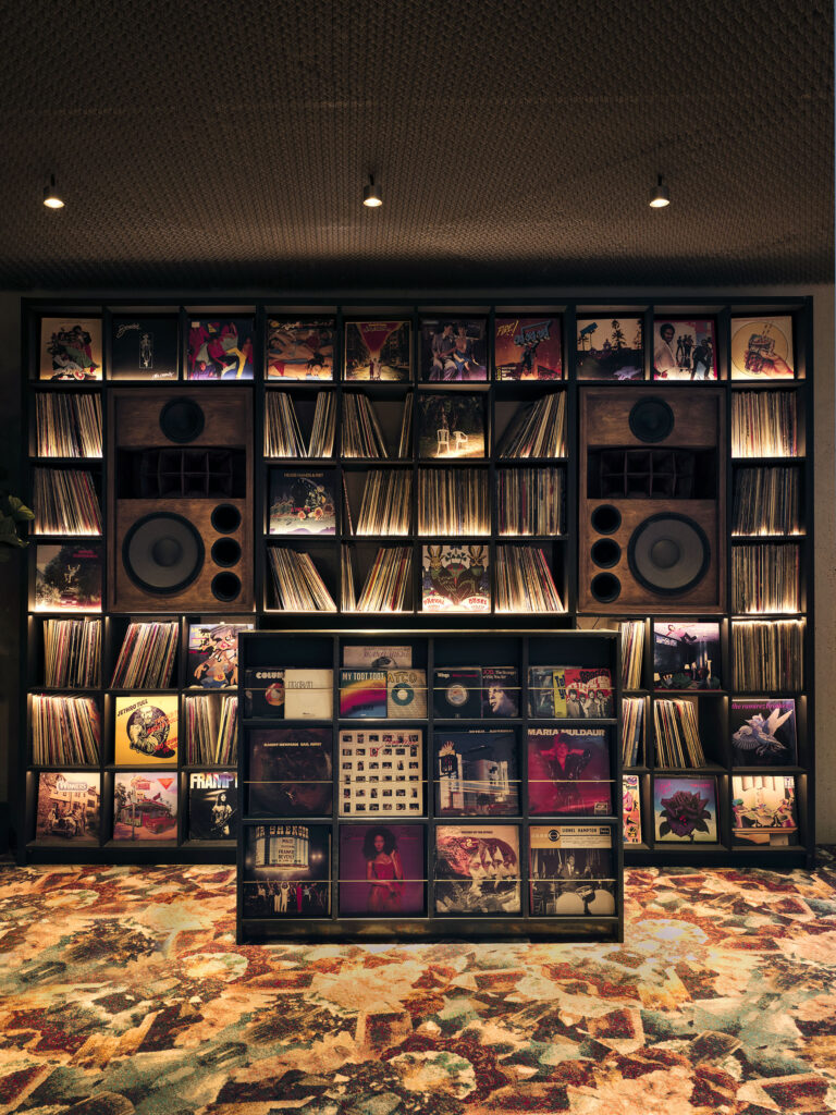 Wall of vinyl records with two large speaker cabinets and a central display in a dim music-store setting.