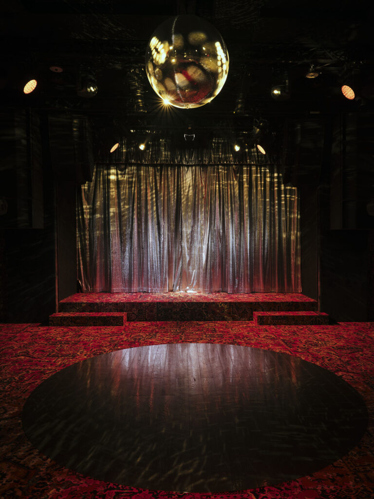 Stage with metallic curtains, disco ball overhead, red patterned carpet, and a circular dark dance floor in front.