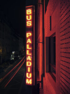Vertical neon sign reading 'BUS PALLADIUM' on a brick building at night, red glow over the street.