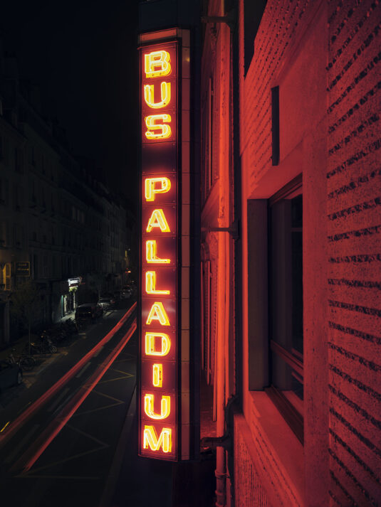 Vertical neon sign reading 'BUS PALLADIUM' on a brick building at night, red glow over the street.