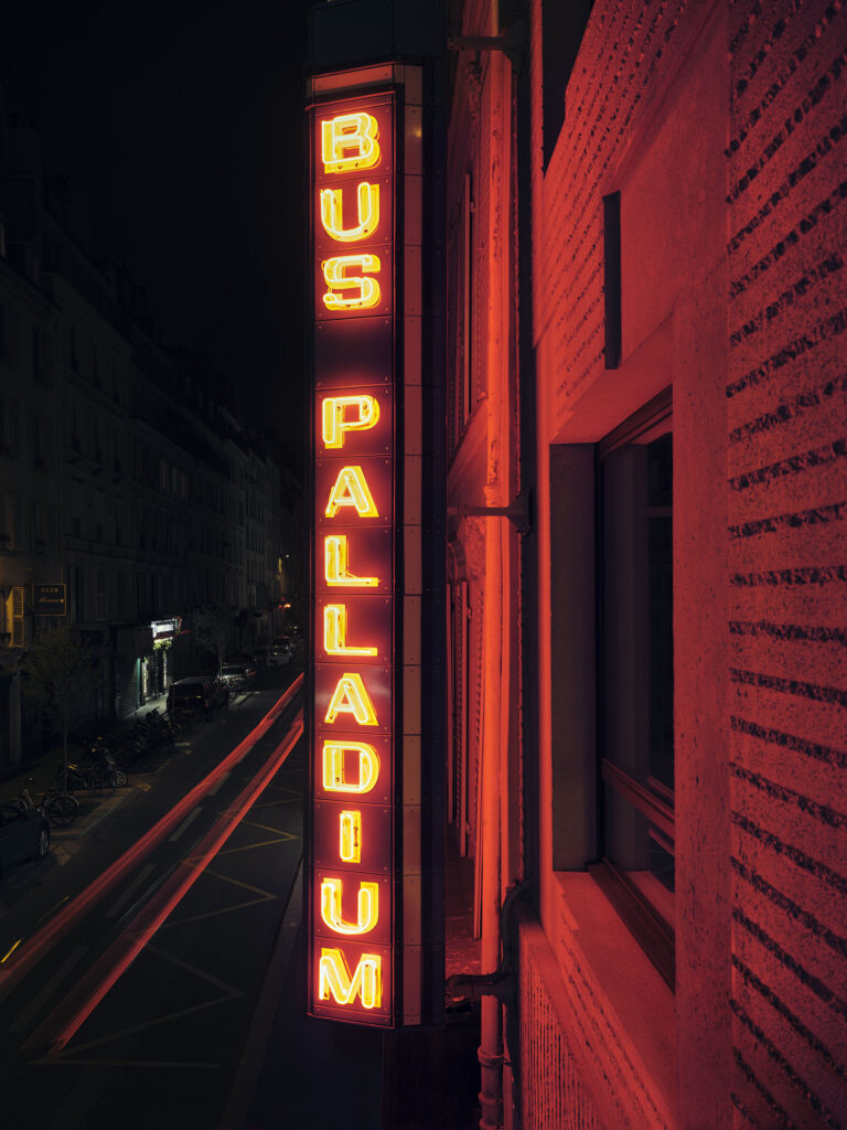 Vertical neon sign reading 'BUS PALLADIUM' on a brick building at night, red glow over the street.