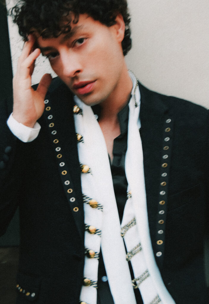 Portrait of a young man with dark, tousled hair wearing a black blazer and a white scarf with gold tassels, looking at the camera while touching his temple.