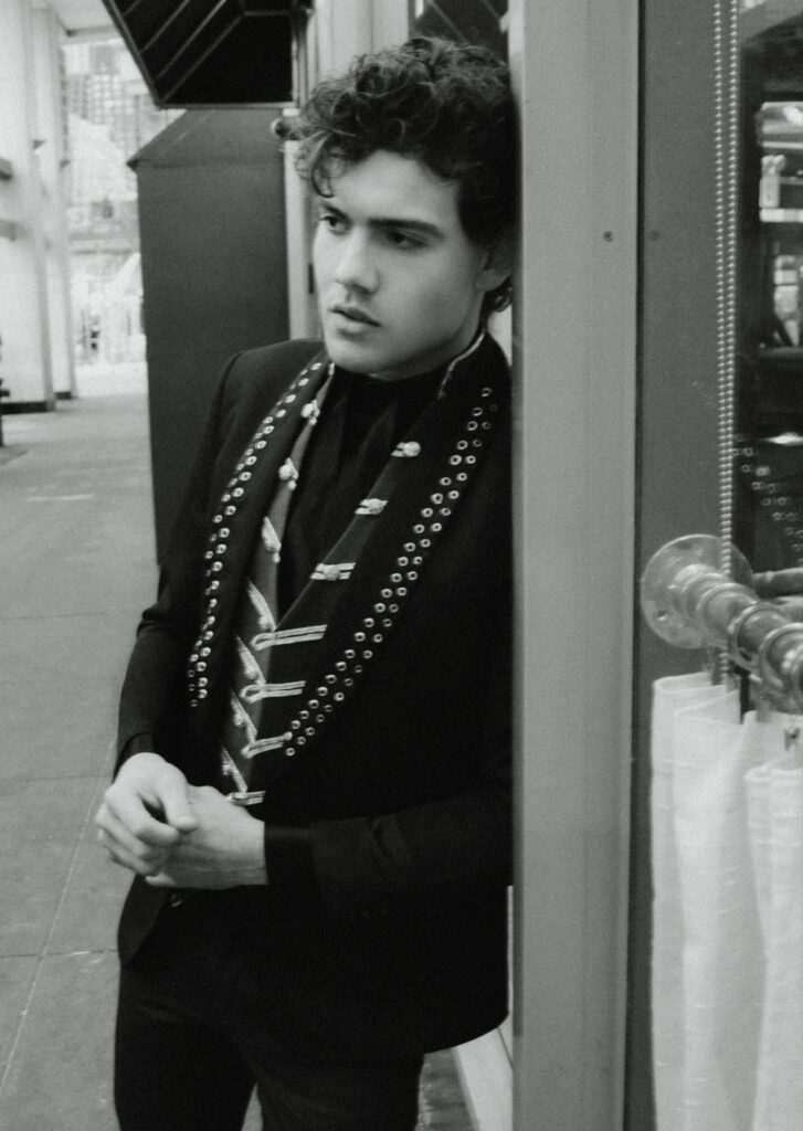 Young man with wavy hair leaning against a storefront, wearing a black blazer and a studded scarf in a city street (black-and-white).