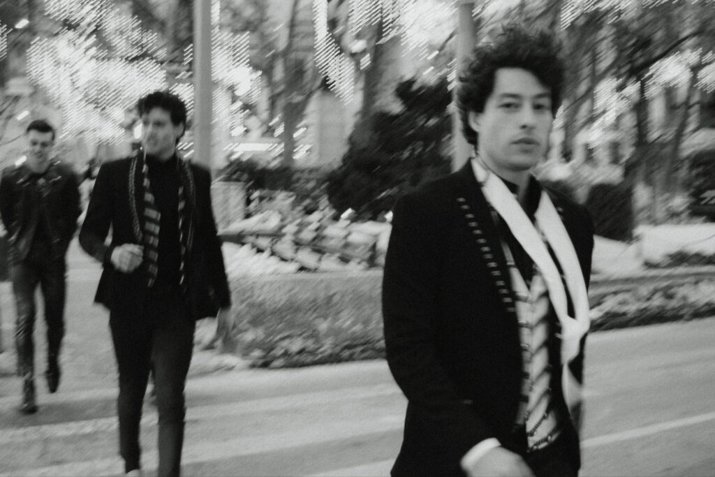 Black-and-white photo of three men walking along a city street at night; the man in the foreground right wears a dark suit and a striped scarf.