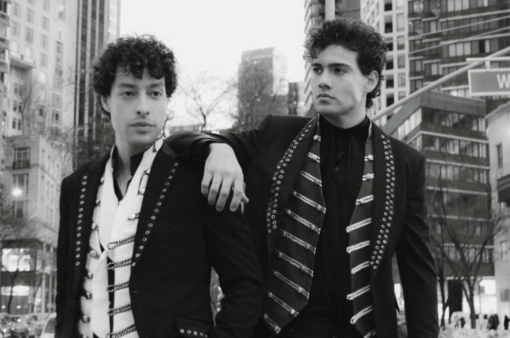 Two stylish men in black suits with decorative scarves standing in a city street, one resting his arm on the other's shoulder as they pose for a photo.