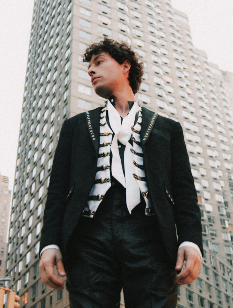Young man in a black military-style jacket with gold braids, in an urban cityscape.