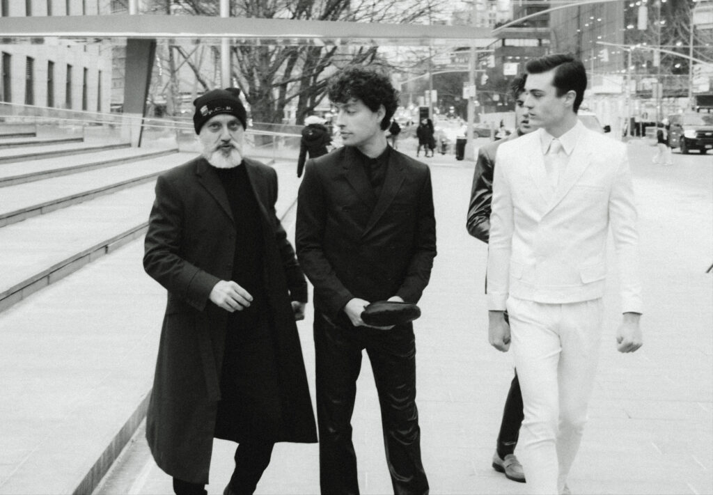 Three men walk together on a city sidewalk; one in a long dark coat and beanie with a white beard, another in a black suit holding a hat, the third in a white suit.
