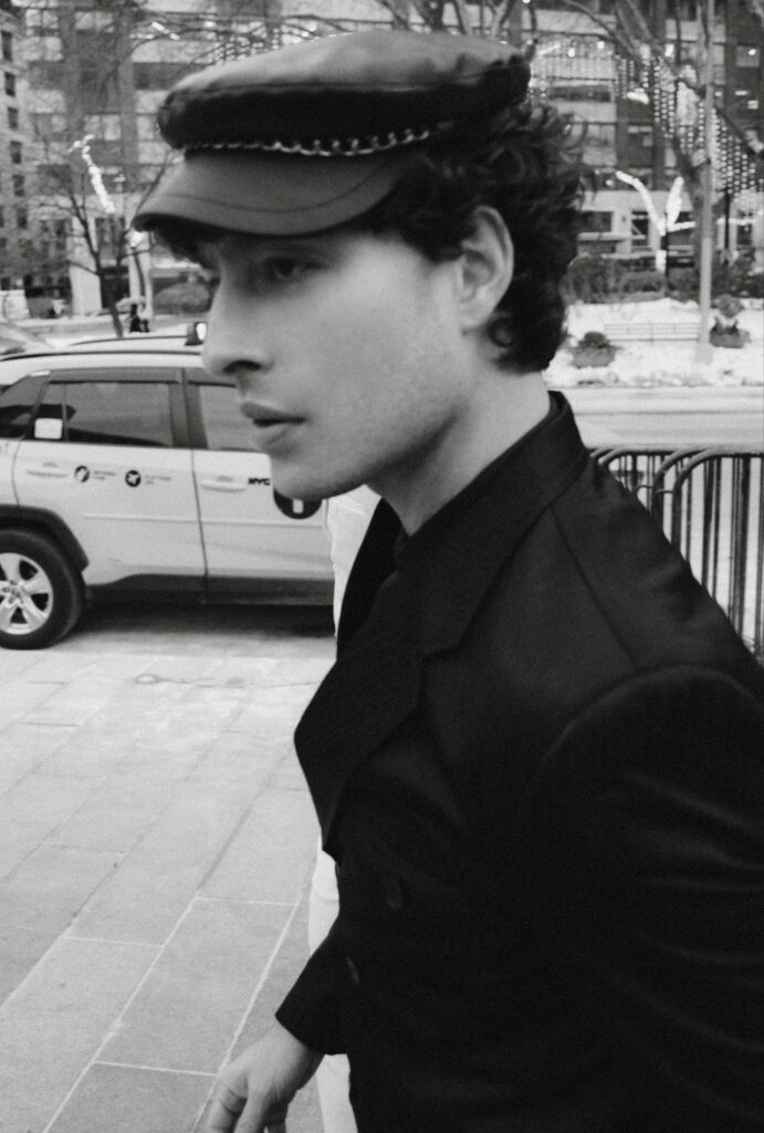 Side-profile of a young man in a dark coat wearing a chain-trimmed cap, standing on a city sidewalk with a taxi in the background.
