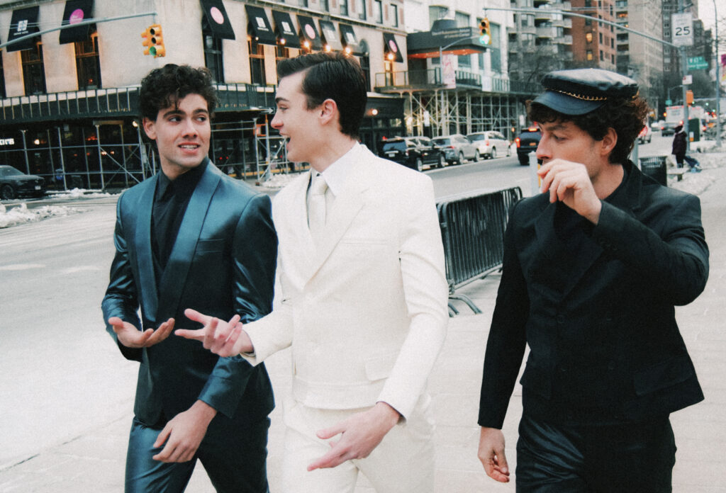 Walk This Way cover story with Three men in stylish suits walking and talking on a city sidewalk with traffic and buildings in the background.