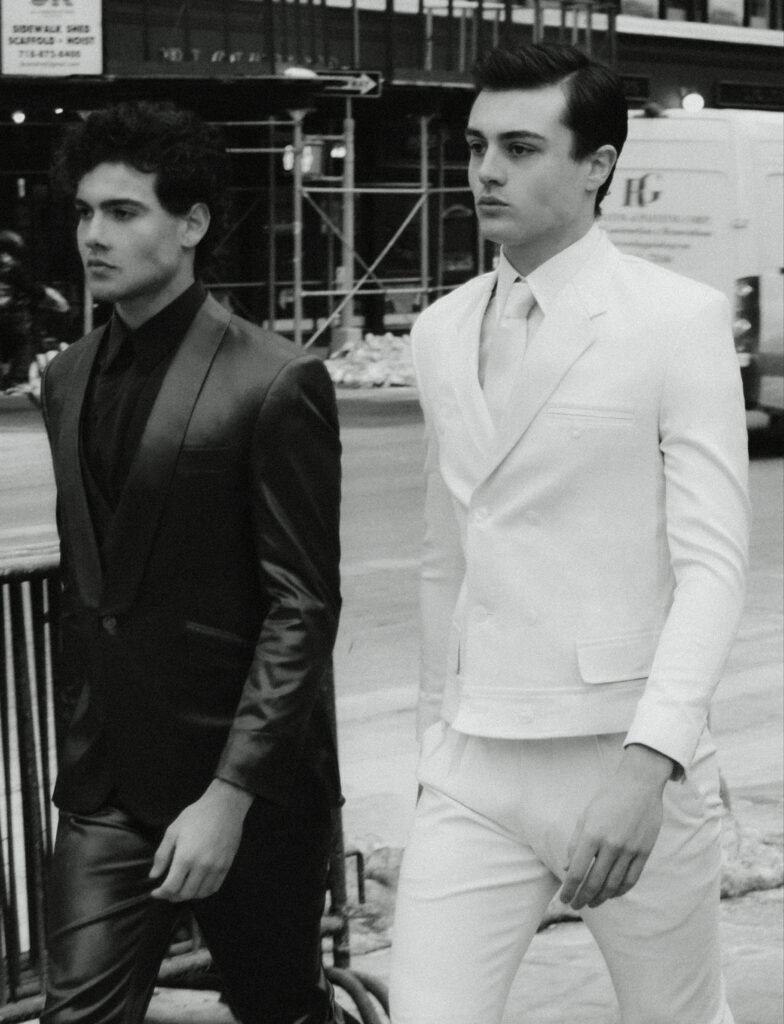 Two men in formal suits walk down a city street—one in a black suit, the other in a white suit—with pedestrians and scaffolding in the background.