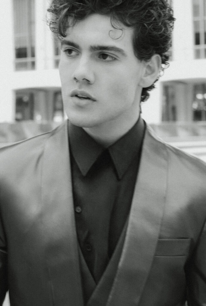 Black-and-white portrait of a young man in a dark shirt and suit vest, looking off to the left, with an urban backdrop.