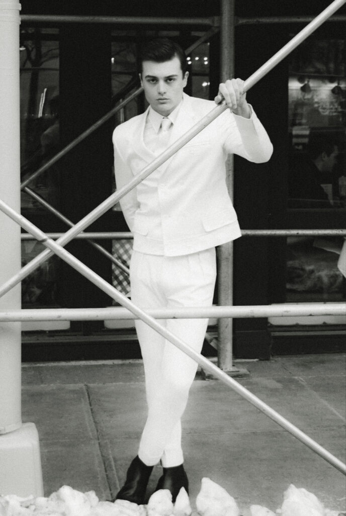Man in a white suit and tie stands beside a diagonal metal railing, leaning and looking at the camera outdoors.