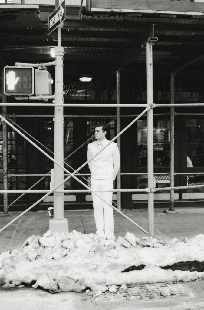 Man in a white suit stands with hands behind his back on a snowy city sidewalk behind scaffolding and construction poles.