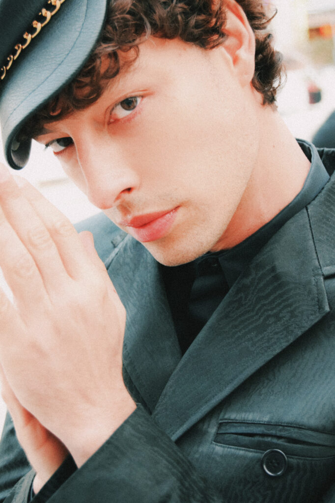 Portrait of a young man in a dark blazer and hat, hands pressed softly together near his face as he gazes at the camera.