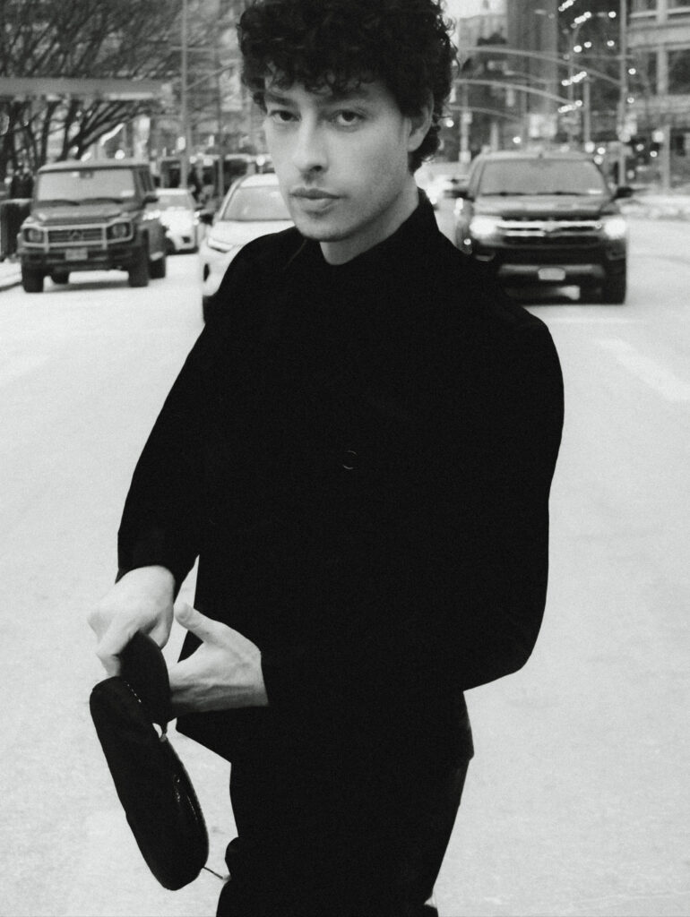 Black-and-white portrait of a young man in a black turtleneck on a city street, adjusting a hat as cars pass in the background.