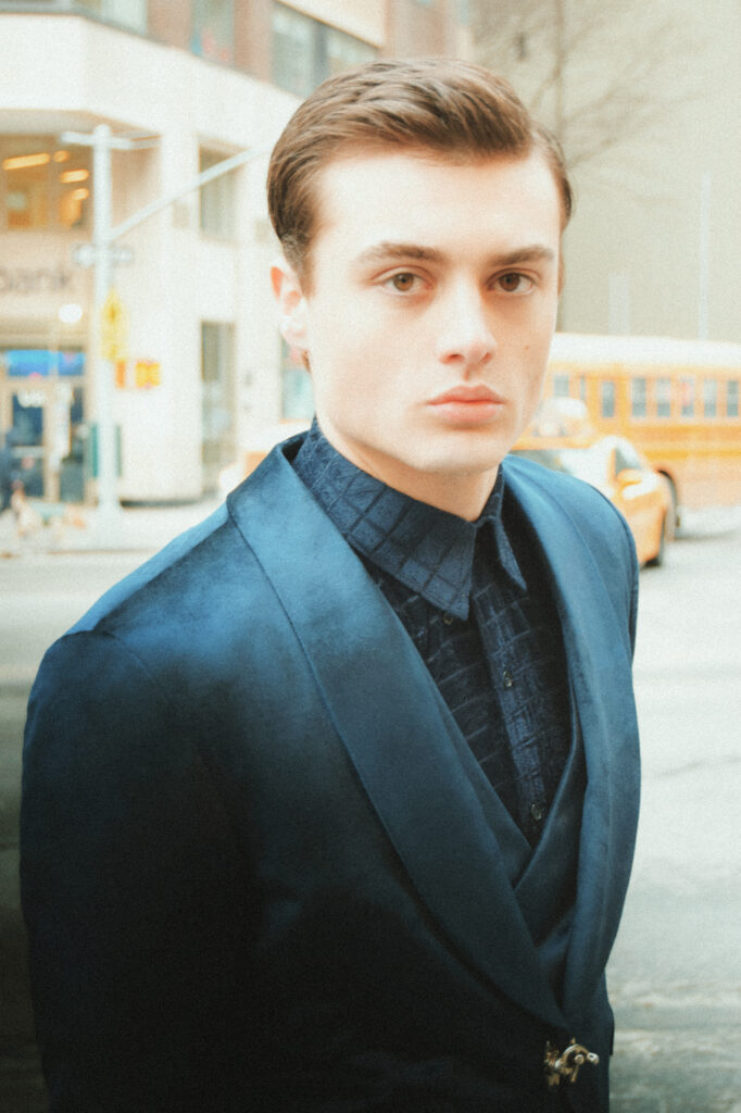 Portrait of a young man in a navy suit jacket on a city street, looking at the camera with a neutral expression behind blurred urban background