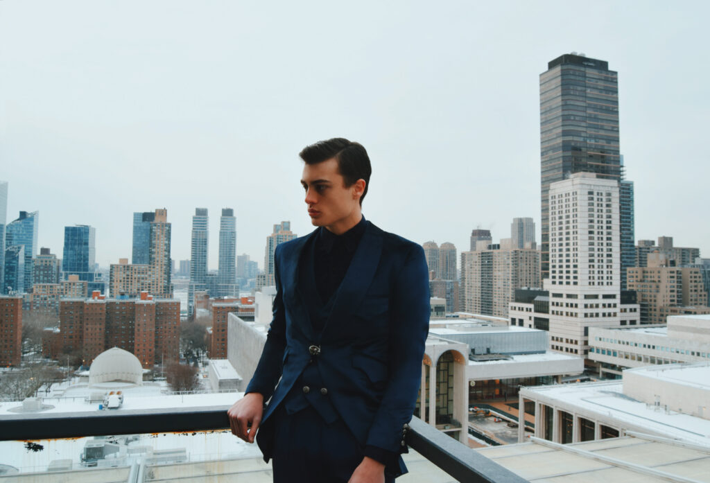 Man in a dark blue suit leaning on a railing on a rooftop with a city skyline in the background, looking to the side.
