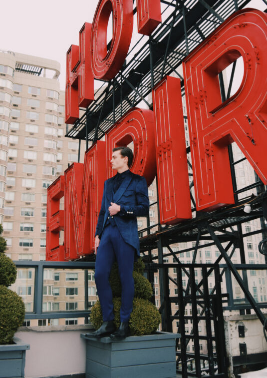 Man in a navy suit standing on a rooftop next to a large red neon sign with city buildings in the background.