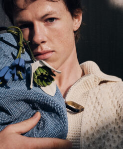 Close-up of Milo Callaghan partially visible face with freckles, wearing a textured blue garment and a beige knit jacket, holding a beaded accessory.
