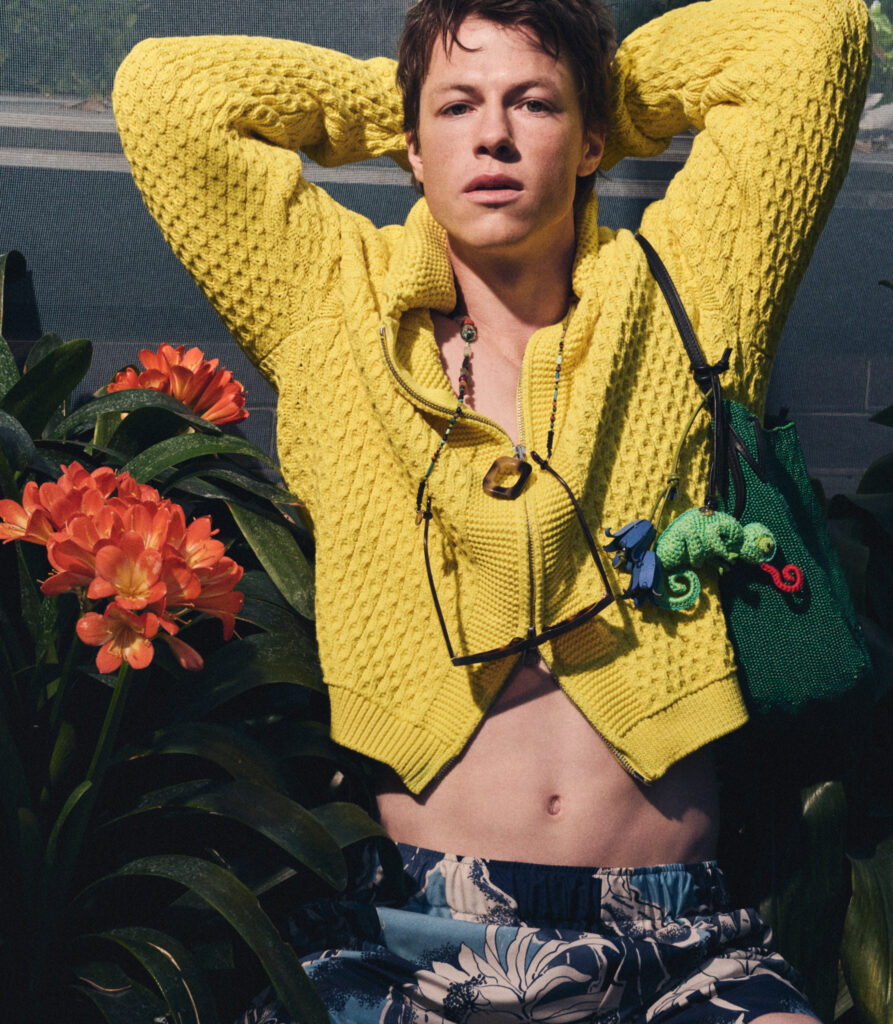 Milo Callaghan in a bright yellow knitted cardigan relaxing among tropical plants, hands behind head, sunglasses hanging from a beaded necklace.