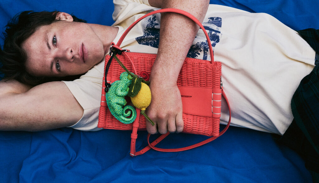 Person lying on a blue surface, holding a red woven handbag with a green/yellow crocheted accessory attached.