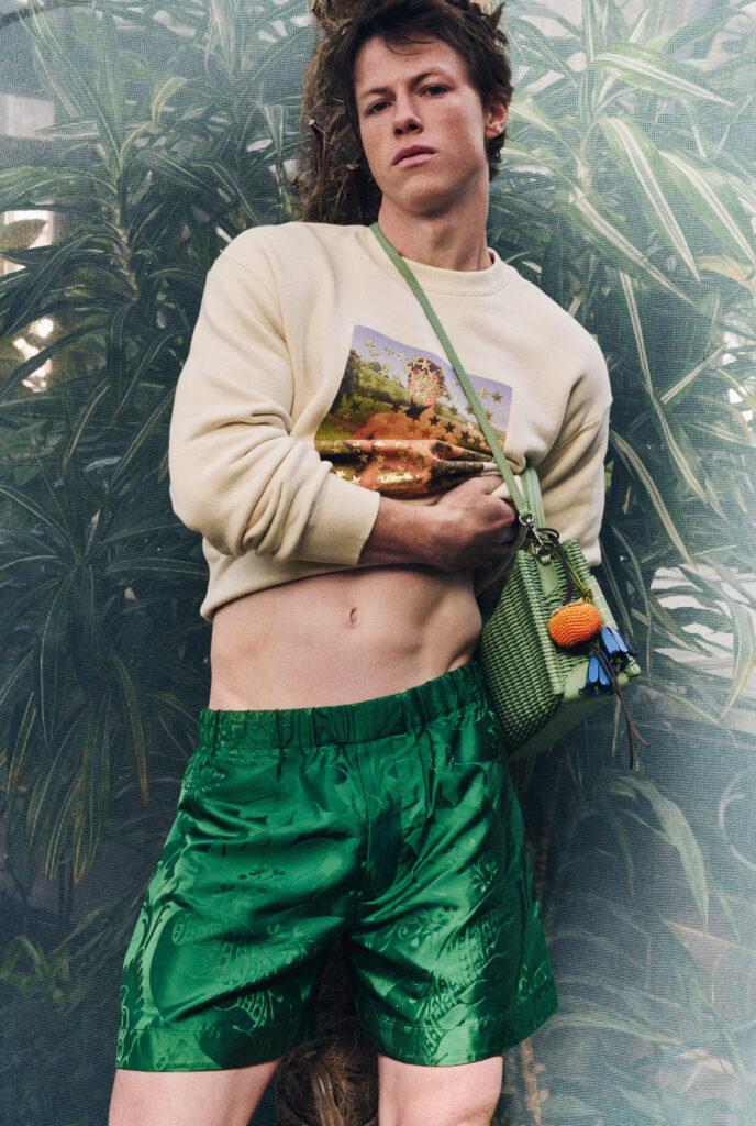 Milo Callaghan posing in a beige sweatshirt and green patterned shorts, holding a green bag, against a backdrop of tropical plants.