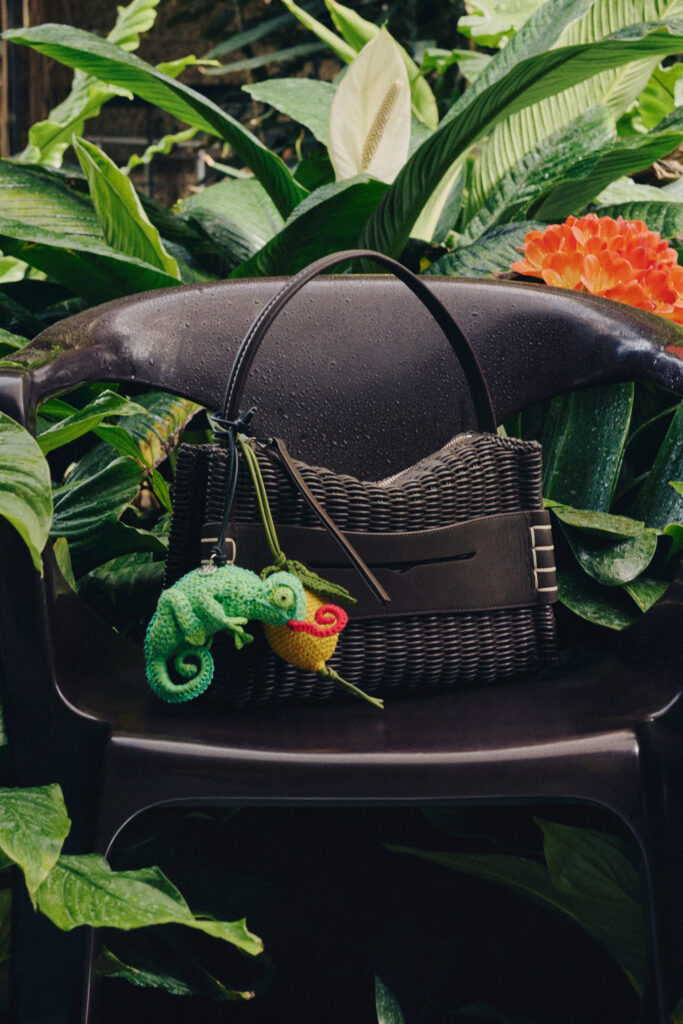 Black helmet resting on a woven basket outdoors, surrounded by large green tropical leaves and an orange flower, with a green knitted octopus hanging from the basket.