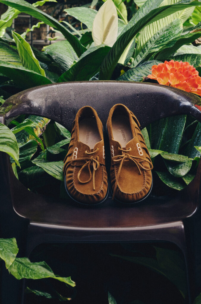 Pair of brown suede moccasin shoes resting on a black chair outdoors, surrounded by lush green tropical plants and an orange flower on the right.