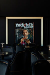 Dark lounge with a large framed poster depicting a man holding a cocktail; black velvet seating and candlelit tables surround it.