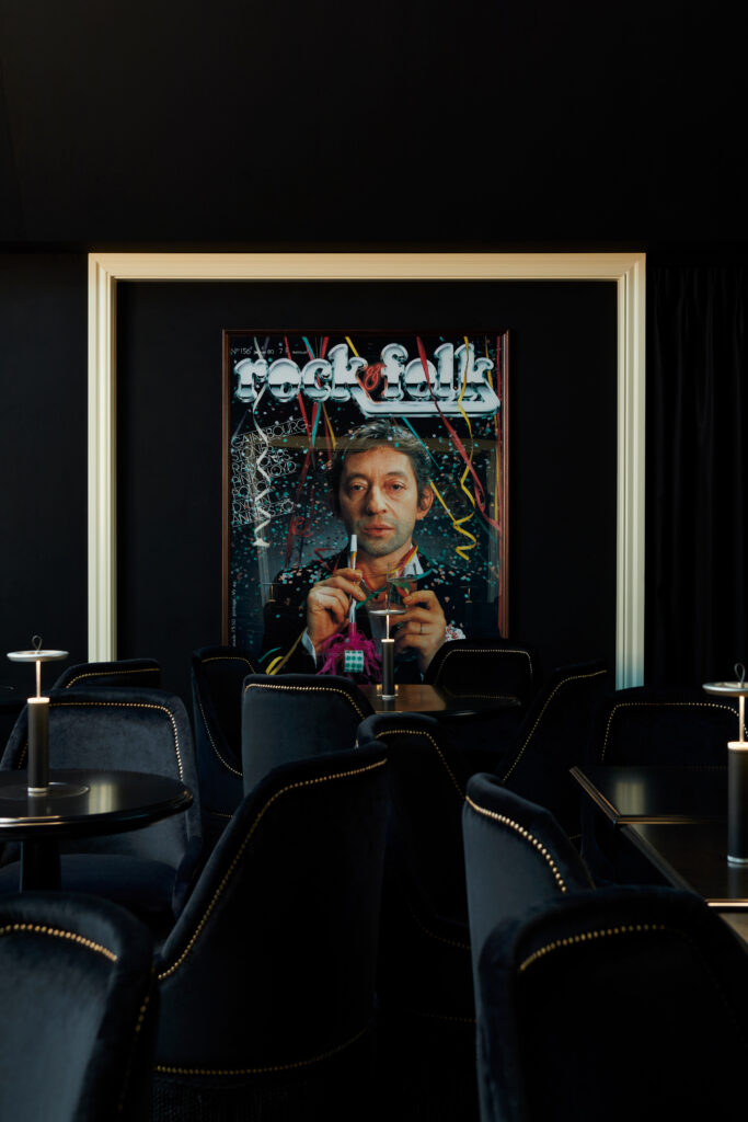 Dark lounge with a large framed poster depicting a man holding a cocktail; black velvet seating and candlelit tables surround it.